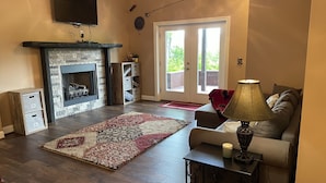 A living room with a fireplace, a sofa, a coffee table with a lamp, and a door leading outside. - Spacious Smoky Mountain Getaway - Sleeps 10, Game Room, Firepit & Views (Maggie Valley)