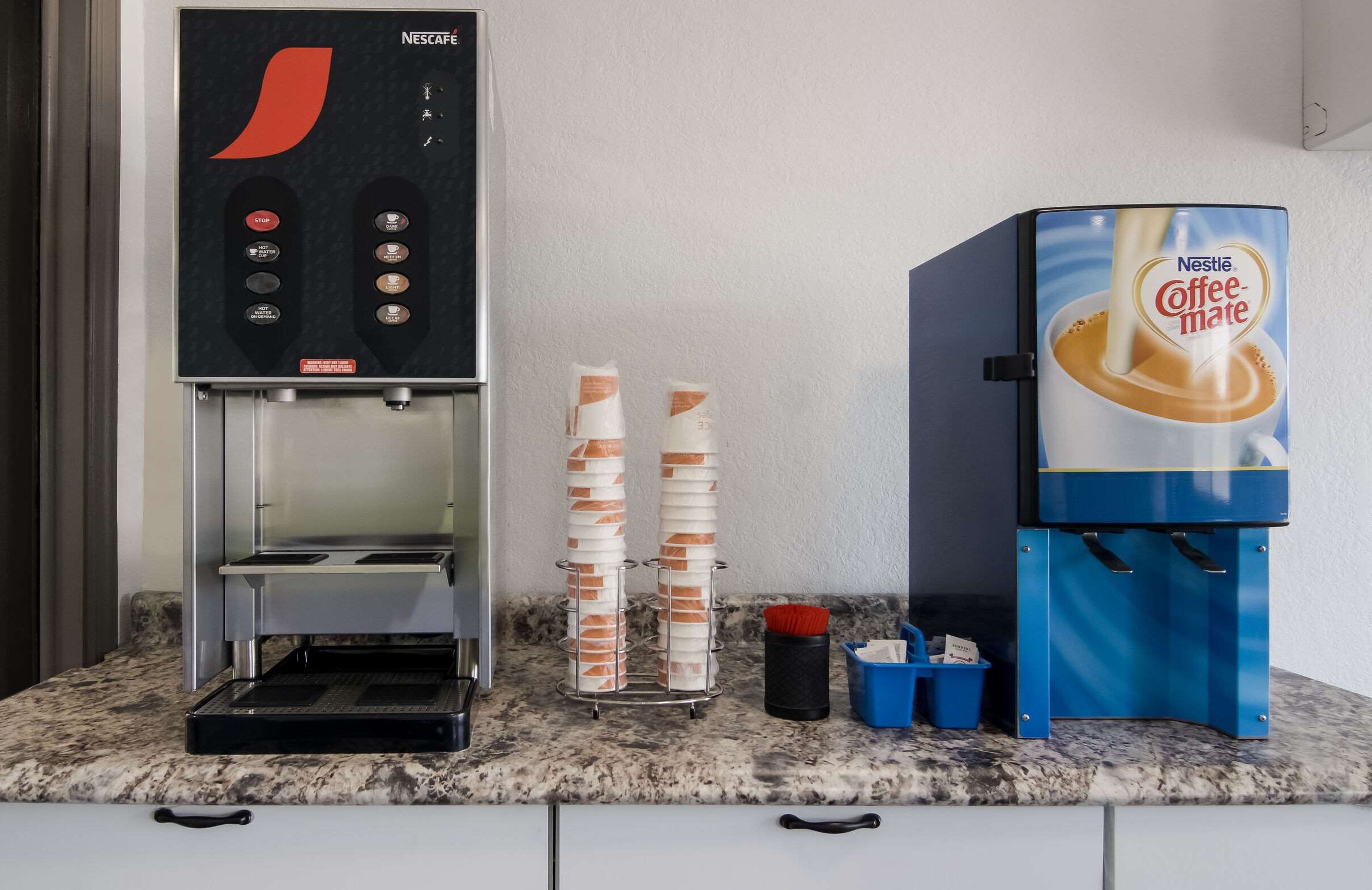 Una cafetera con un dispensador de Nestle Coffee-Mate y una pila de vasos desechables.