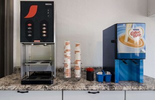 Una cafetera con un dispensador de Nestle Coffee-Mate y una pila de vasos desechables.