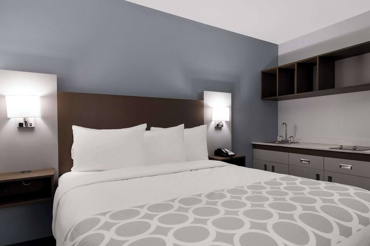 A hotel room with a bed, bedside tables, a nightstand, a wall-mounted shelf, and a small kitchenette area.