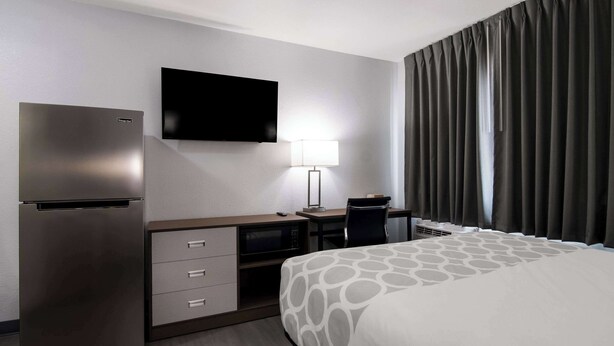 A hotel room with a bed, a desk with a chair, a flat-screen TV, a refrigerator, a lamp, and a window with curtains.