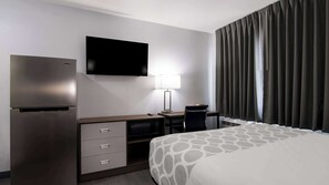 A hotel room with a bed, a desk with a chair, a flat-screen TV, a refrigerator, a lamp, and a window with curtains.