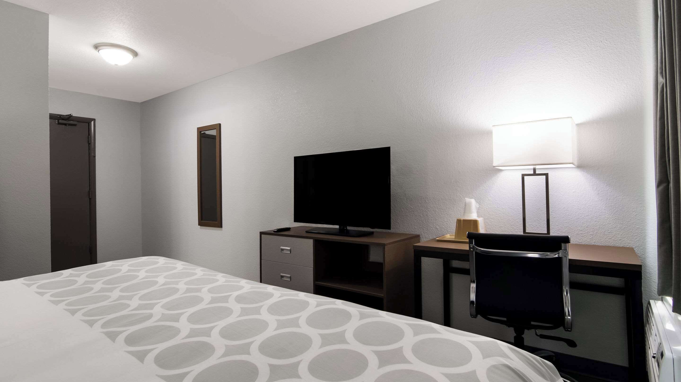 A hotel room with a bed, a desk with a television, a chair, a lamp, and a door.