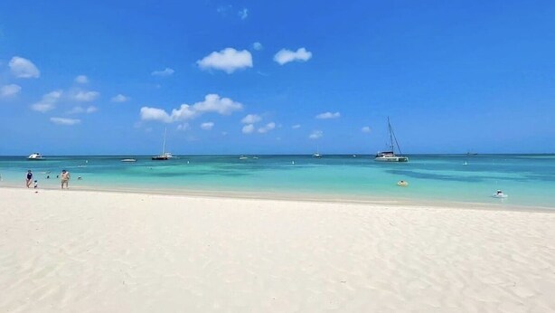 Ein Strand mit klarem türkisblauem Wasser, einem sandigen Ufer und mehreren Segelbooten in der Ferne.