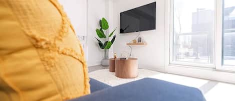 Een moderne woonkamer met een groot raam, een flatscreen televisie, een blauwe bank en een houten bijzettafeltje met een plant.