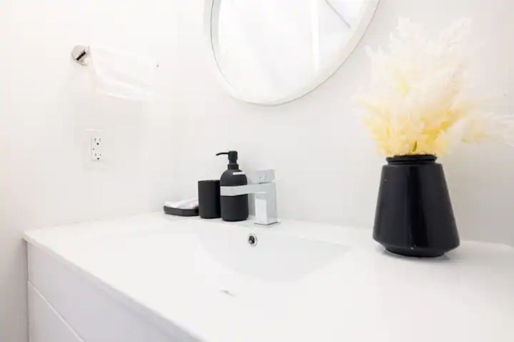 Un tocador blanco con un dispensador y bomba de jabón negros, un jarrón negro con flores blancas, y un espejo redondo en la pared.
