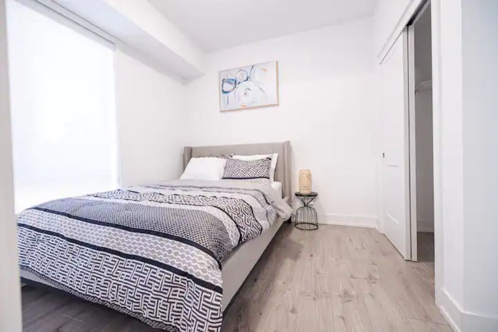 A bedroom with a bed, a bedside table with a lamp, a painting on the wall, and a door.