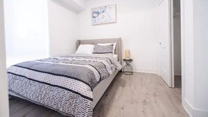 A bedroom with a bed, a bedside table with a lamp, a painting on the wall, and a door.