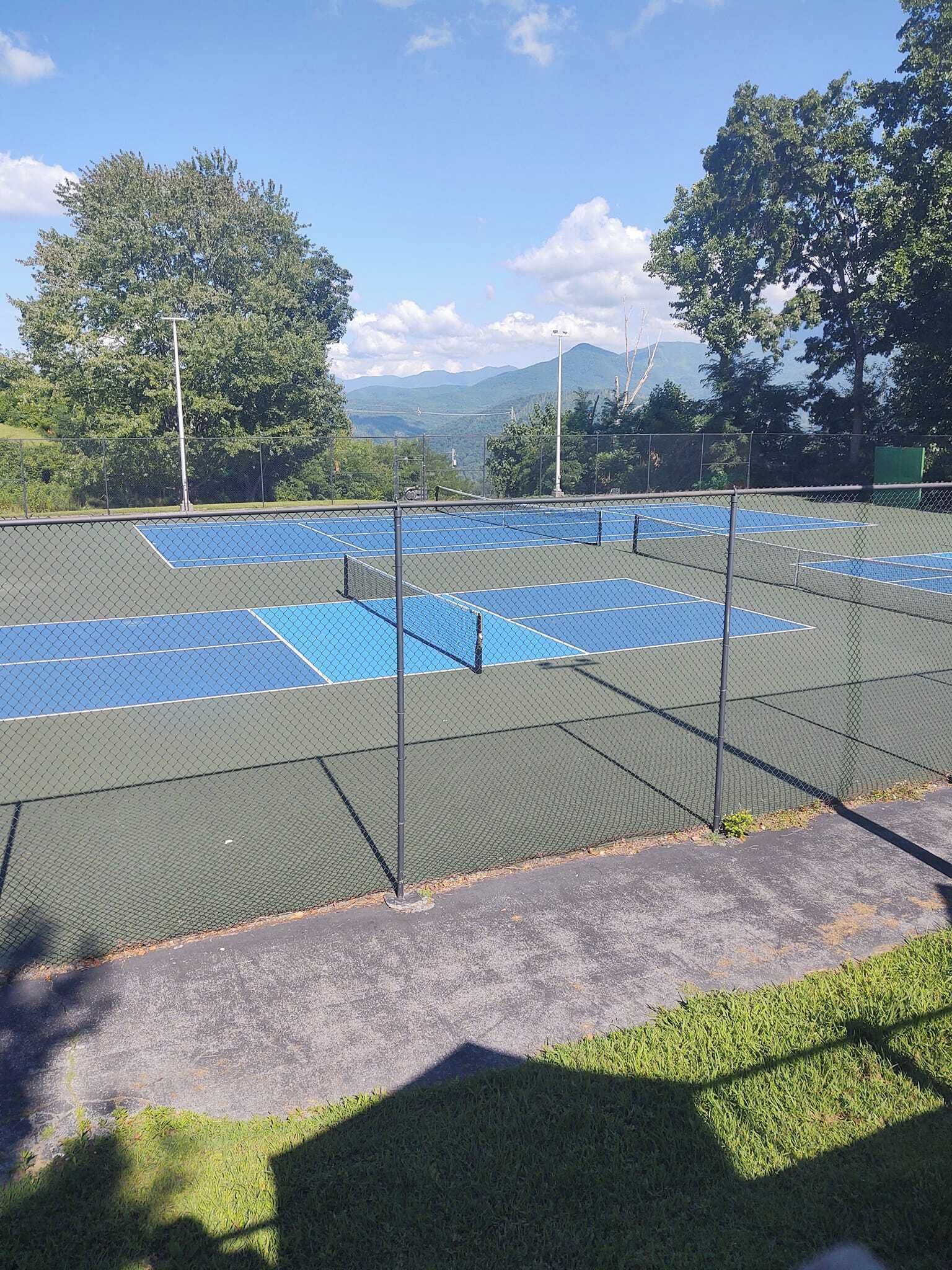 Un court de tennis en plein air, entouré d’une clôture en treillis métallique, bordé d’arbres et avec une chaîne de montagnes en arrière-plan.