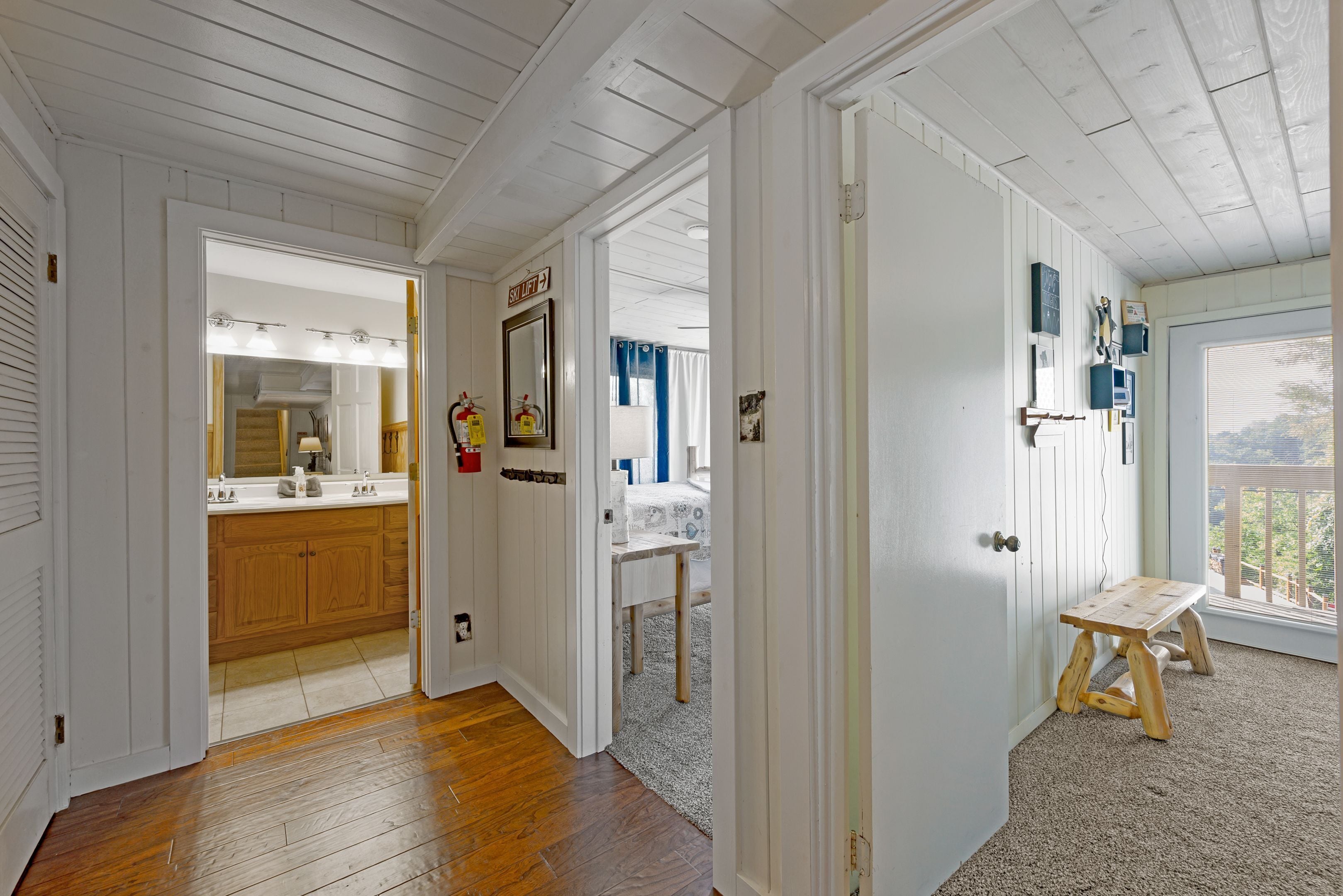 A hallway with wooden flooring, a mirror with lights, a sink, and a small wooden stool.