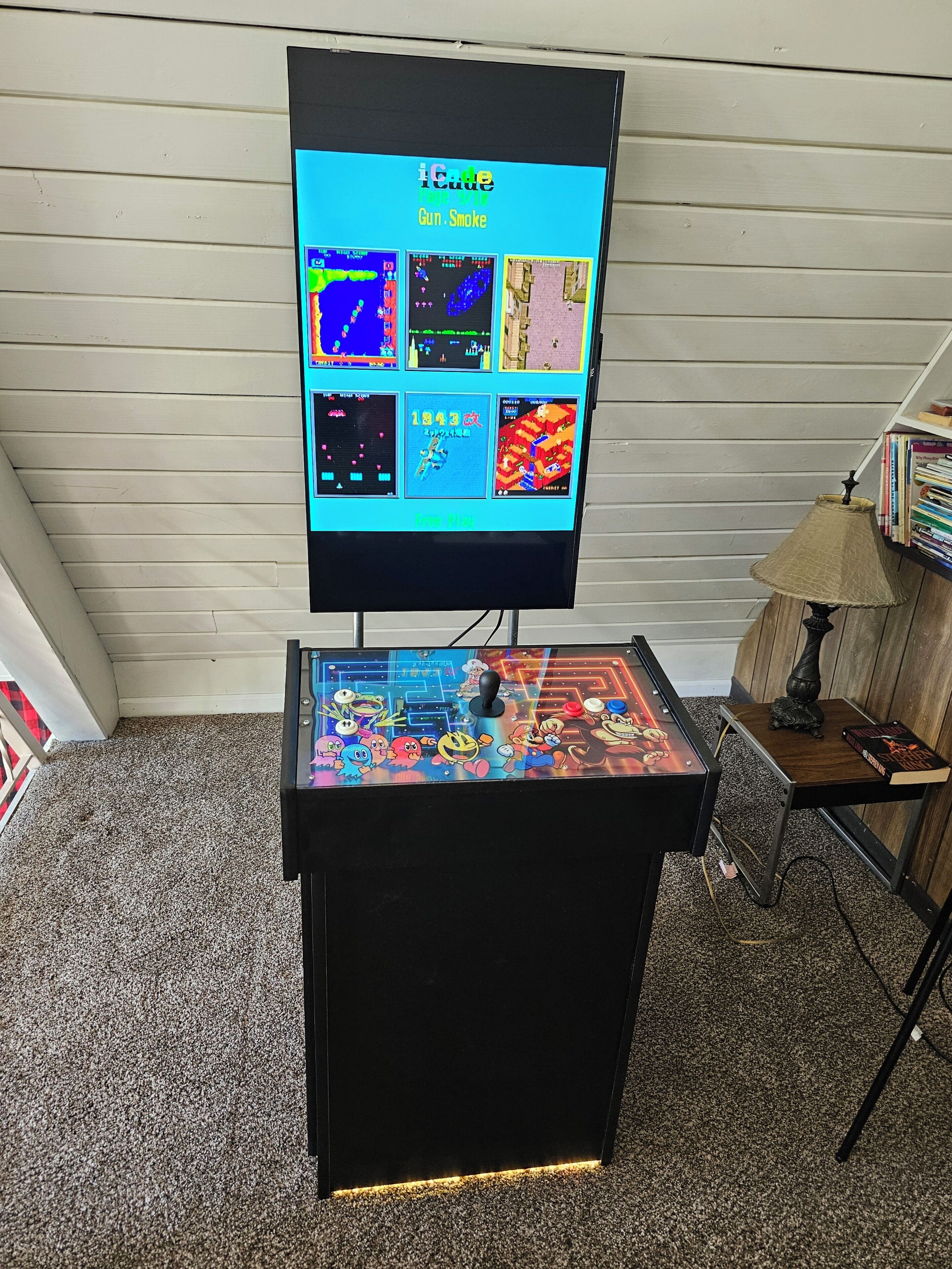 Une salle d’arcade de jeux vidéo à l’ancienne, avec une sélection de jeux affichés sur un écran, un joystick et une petite table avec une lampe.