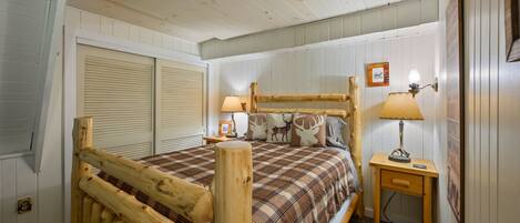 A wooden bed with a plaid bedspread, two bedside lamps, and a small wooden nightstand with a lamp.