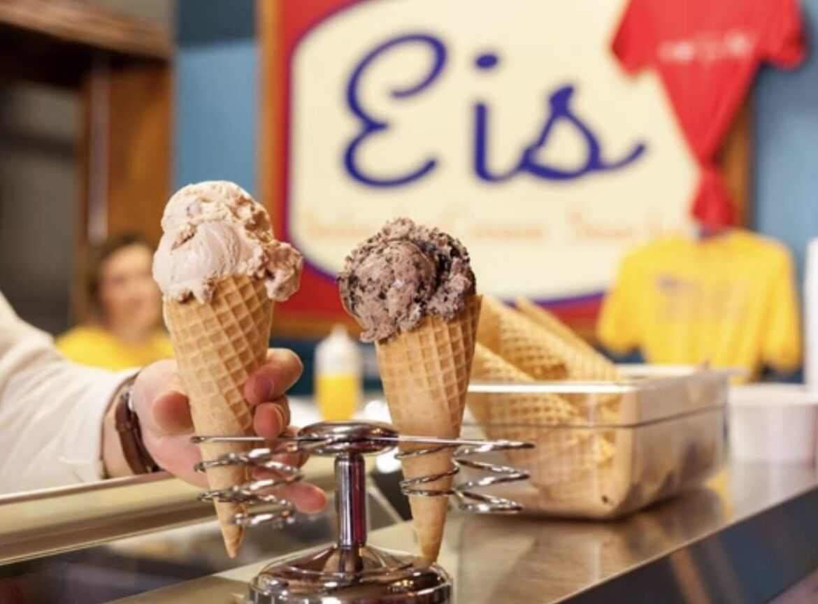 Dos helados en conos sobre un soporte metálico, con un fondo desenfocado que muestra un letrero con la palabra 'Eis'.