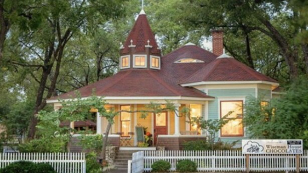 Een huis in Victoriaanse stijl met een opvallende toren, een veranda met een groene deur en een bord waarop Wiseman House Chocolates staat aangeboden.