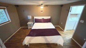 Ein Schlafzimmer mit Bett, Nachttisch, Lampe, Deckenventilator und Blick auf einen Waschbereich.
