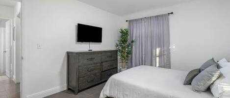 Ein Schlafzimmer mit Bett, Kommode, Fernseher und einem Fenster mit Vorhängen.