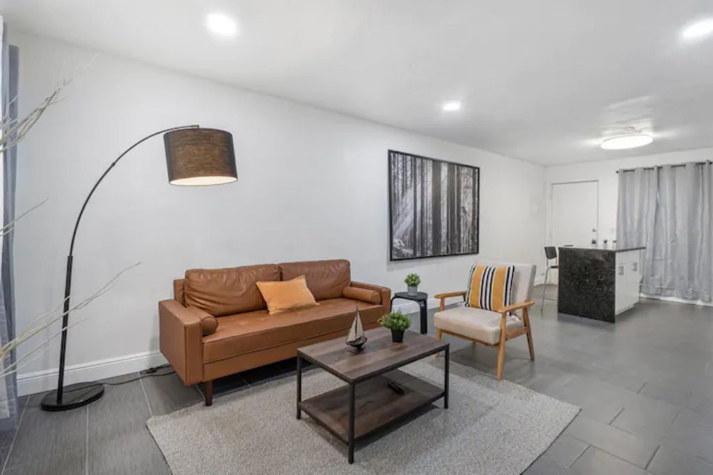 A modern living room with a brown leather sofa, a wooden coffee table, and a floor lamp. There is a painting on the wall and a small plant on the coffee table.