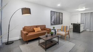 Una sala de estar moderna con un sofá de cuero marrón, una mesa de centro de madera y una lámpara de pie. Hay un cuadro en la pared y una planta pequeña sobre la mesa de centro.