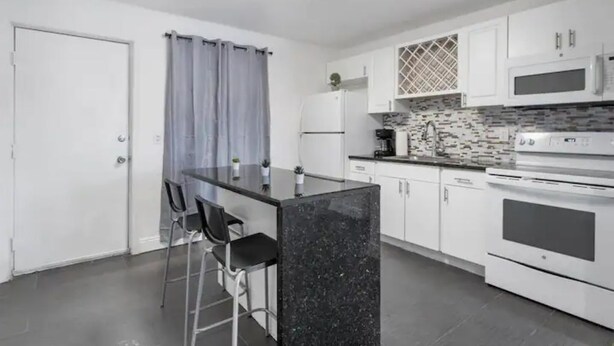 Una cocina compacta con armarios blancos, una isla de mármol negro y un pequeño comedor con dos sillas.