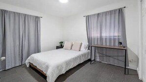 Un dormitorio con cama, cortinas, una mesita de noche y una mesita auxiliar.