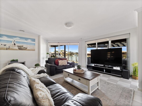 Una sala de estar moderna con un sofá de cuero, una mesa de centro y un televisor de pantalla plana. Una ventana grande ofrece vistas al mar y a barcos.