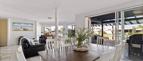A spacious living area with a wooden dining table, white chairs, and a large window overlooking a pool and outdoor seating area.