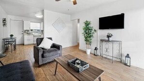 Een moderne woonkamer met een grijze bank, een houten salontafel, een plafondventilator, een aan de muur bevestigde televisie en een potplant.
