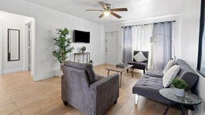 Een moderne woonkamer met een grijze bank, een grijze fauteuil, een klein rond tafeltje, een plafondventilator, een aan de muur bevestigde televisie en een potplant.