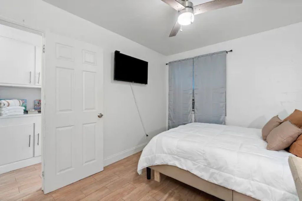 Une chambre à coucher avec un lit, un ventilateur de plafond, un téléviseur fixé au mur et une fenêtre avec un rideau.