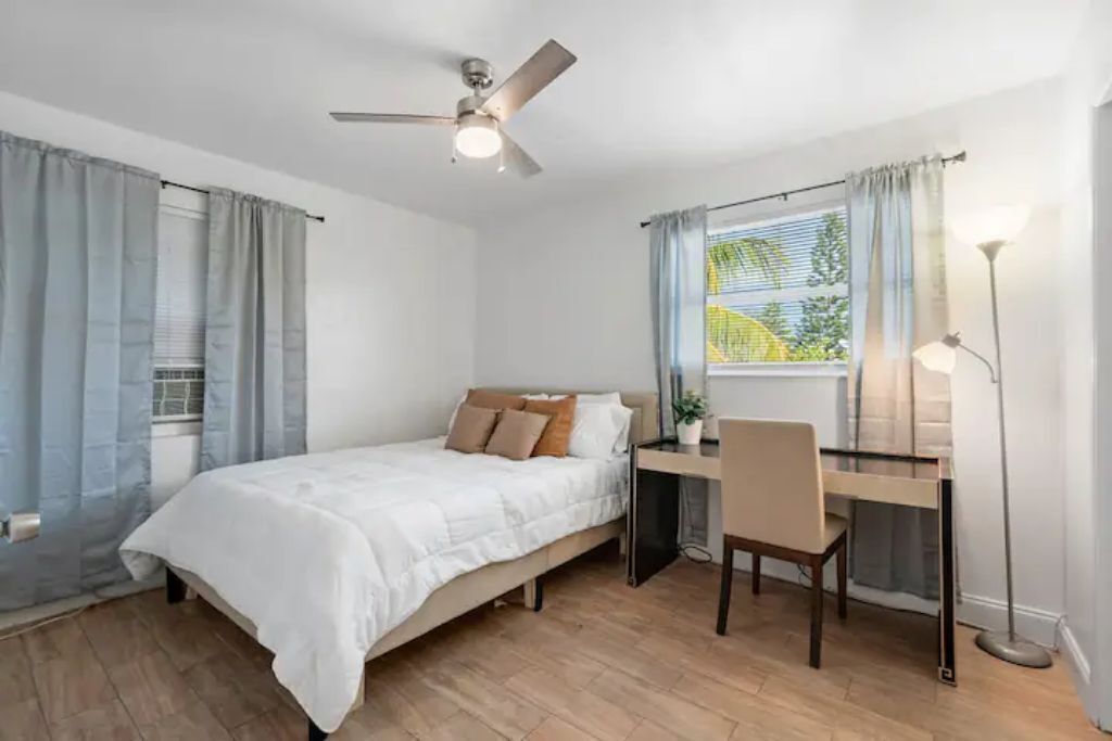 A bedroom with a bed, desk, chair, ceiling fan, and window with curtains.