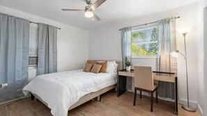 Een slaapkamer met een bed, bureau, stoel, plafondventilator en raam met gordijnen.