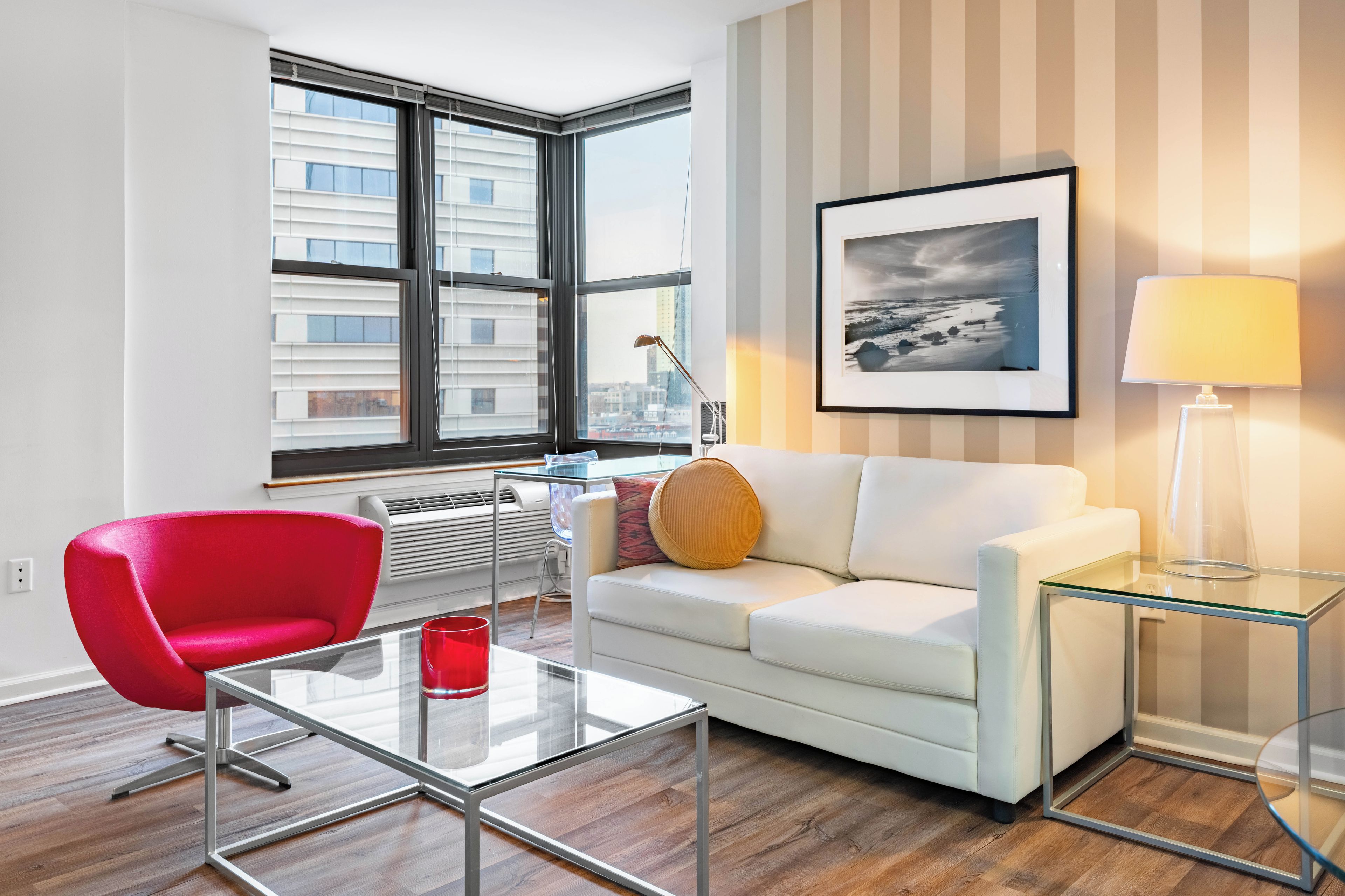 Een moderne woonkamer met een rode fauteuil, een witte bank, een glazen salontafel en een stadszicht door grote ramen.