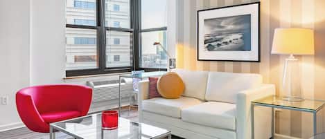 A modern living room with a red chair, white sofa, glass coffee table, and a city view through large windows.