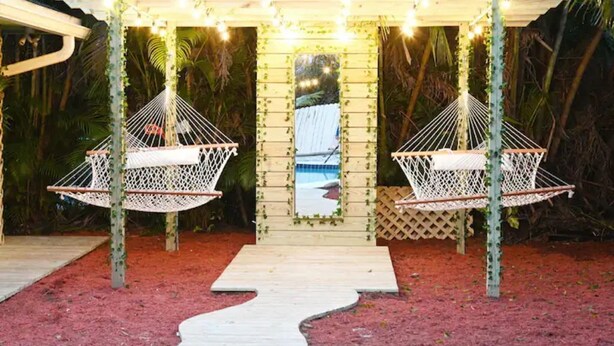 Een houten pergola met twee witte hangmatten, een pad naar een houten terras en een zwembad op de achtergrond.
