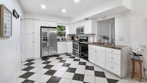 Cozinha moderna com armários brancos, piso xadrez preto e branco, eletrodomésticos de aço inoxidável e um pequeno banquinho de madeira com uma planta.
