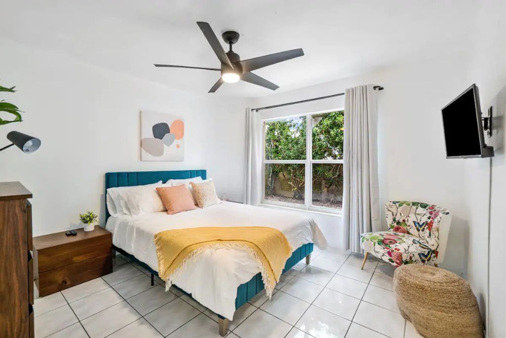 A bedroom with a bed, a ceiling fan, a window with curtains, a TV, and a chair.