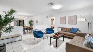 Uma sala de estar moderna com um sofá, duas poltronas azuis, uma mesa de centro e um espaço de jantar com uma mesa e cadeiras.
