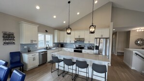Uma cozinha moderna com armários brancos, uma ilha grande com banquetas, eletrodomésticos de aço inoxidável e iluminação pendente.