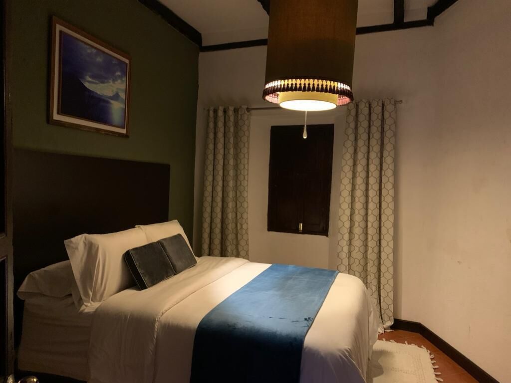 Habitación de hotel con cama, lámpara colgante, cortinas en la ventana y un cuadro en la pared.
