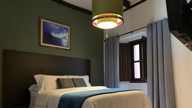 Un lit bien fait, recouvert d’un plaid bleu, avec un tableau encadré accroché au mur et une lampe suspendue au-dessus.