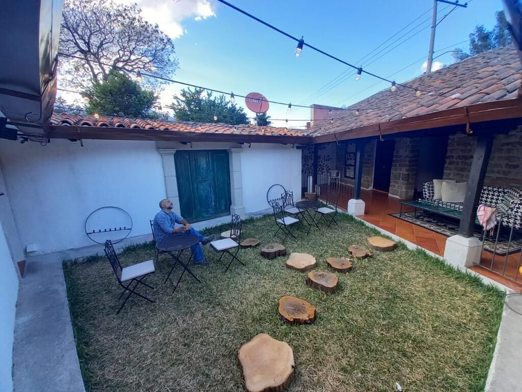 Un patio con pared blanca, puerta verde y techo de tejas. Varias sillas y mesas están dispuestas sobre el césped.