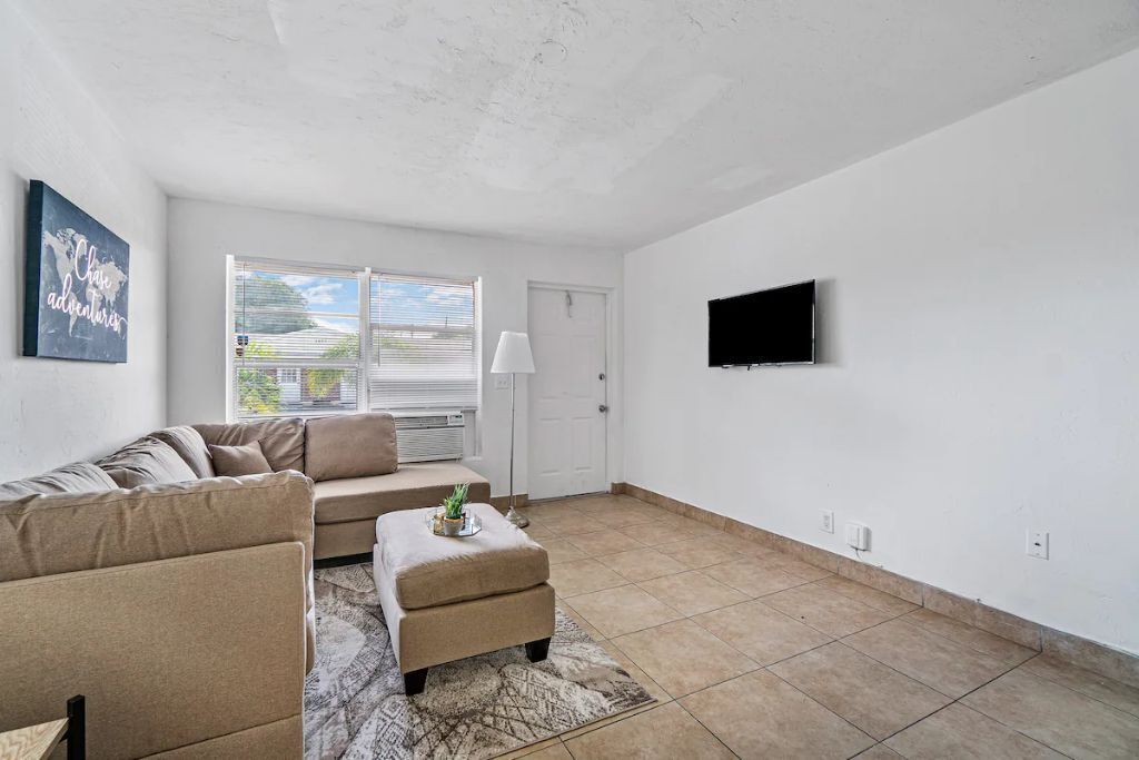 A living room with a beige sofa, a small ottoman, a flat-screen TV mounted on the wall, and a window with blinds.