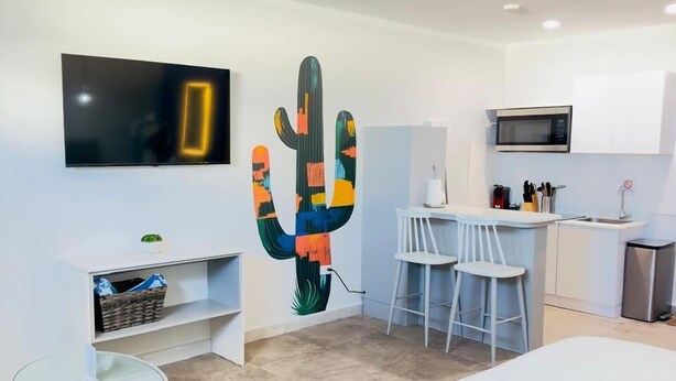 Une chambre d’hôtel moderne équipée d’un lit, d’une t élévision, d’une décoration murale représentant des cactus, d’une petite table ronde et d’une kitchenette.