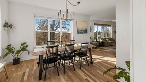 Een eethoek met een houten tafel, zwarte stoelen en een kroonluchter. Grote ramen bieden uitzicht op bomen en er hangt een schilderij aan de muur.