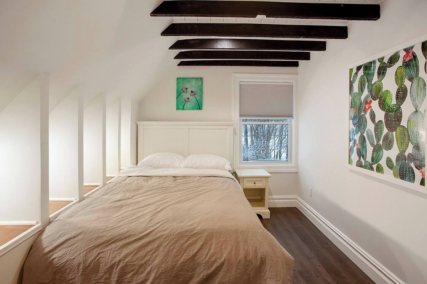 Un dormitorio con cama, mesita de noche, ventana con vistas y una obra de arte con temática de cactus en la pared.