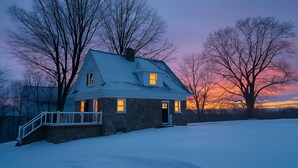 Une maison en pierre avec un toit à pignon, un porche à l’avant et un paysage enneigé.