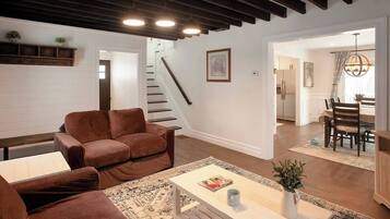 A spacious living room with a brown leather sofa, a white coffee table, and a patterned rug. A staircase leads to an upper floor, and a dining area is visible in the background.