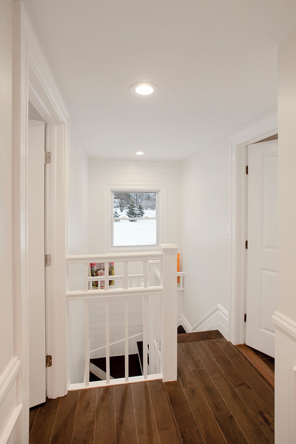 A hallway with wooden flooring, white walls, and a window with a view of trees and snow.