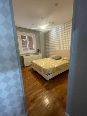 Ein Schlafzimmer mit einem Bett, einem Deckenventilator, einem Fenster mit Jalousien, einem Schminktisch und strukturierter Tapete.