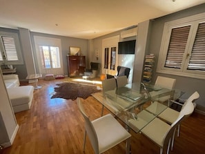 A modern living room with a glass dining table, white chairs, a leather rug, and a fireplace.
